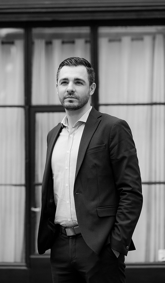 Cette image en noir et blanc montre Michael Stolard, expert comptable à Les Angles, vêtu d'un costume. Il adopte une posture confiante, regardant au loin avec sérieux, reflétant son professionnalisme et son engagement envers ses clients. L'arrière-plan, avec une façade élégante, ajoute à l'ambiance professionnelle. Cette image illustre parfaitement son rôle en tant qu'expert-comptable, offrant des services de qualité aux entreprises locales de Les Angles.
